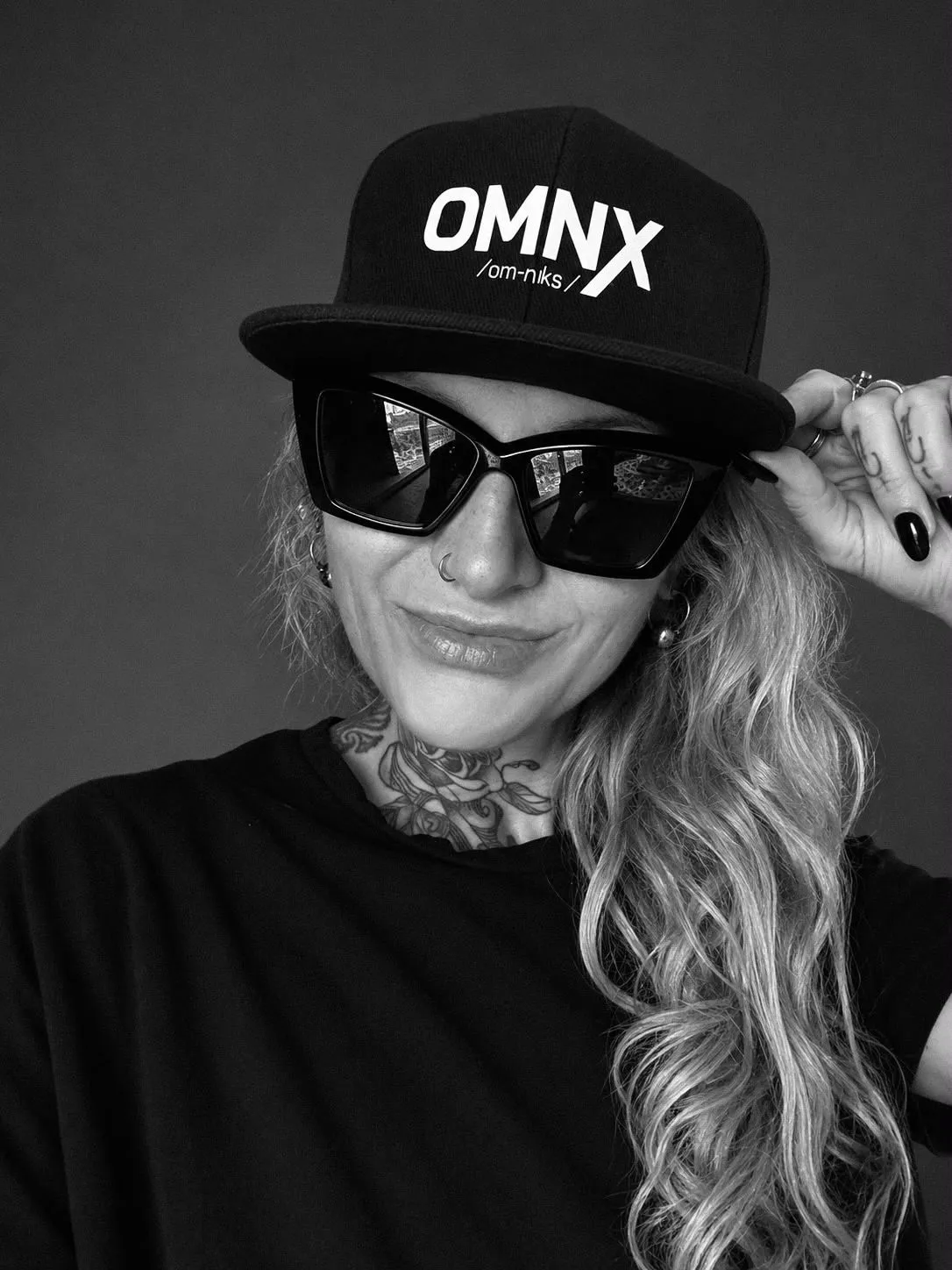 Black and white photograph of a person wearing the cap head on, tilting the flat brim up with one hand, OMNX and the pronunciation /om-niks/ across the front panels