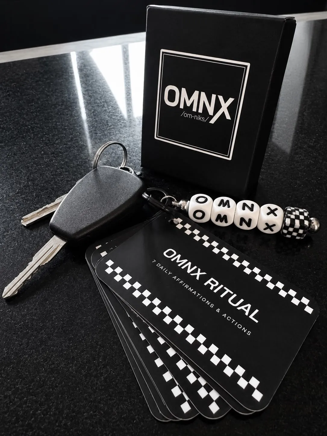 The card set fanned open on a black worktop beside its OMNX gift box and a set of keys, the beaded OMNX charm threaded onto the ring, the top card reading OMNX RITUAL over 7 DAILY AFFIRMATIONS & ACTIONS