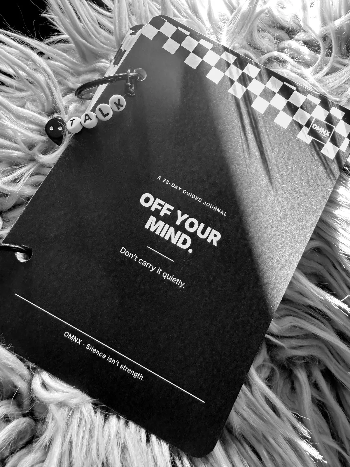 The black cover reading A 28-DAY GUIDED JOURNAL, OFF YOUR MIND. and Don't carry it quietly., a chequered band across the top corner and a beaded TALK charm on the binding ring, lying on a shaggy rug