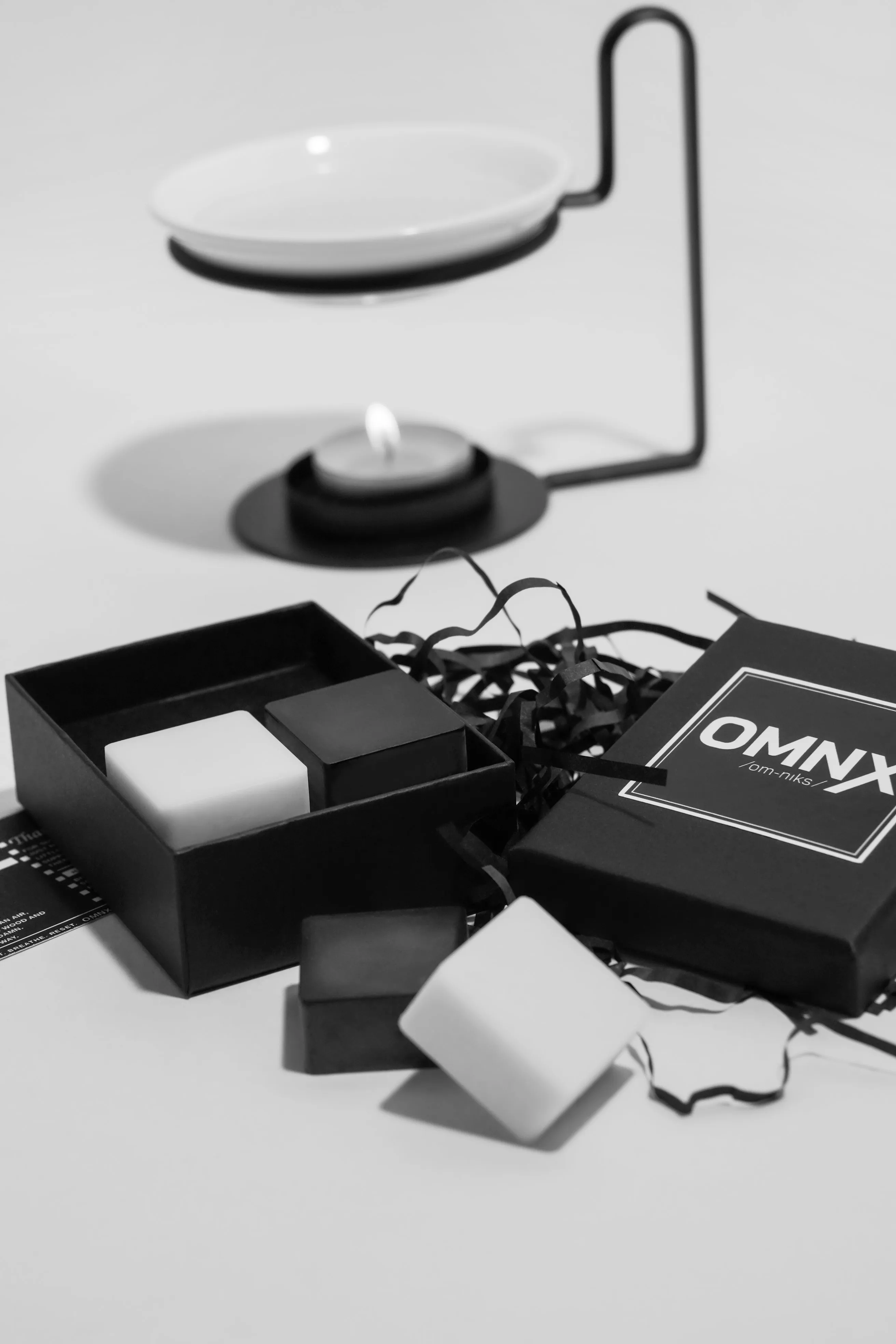 A black and white photograph of a ceramic burner, its dish empty and a tea light burning beneath it, set behind an open Drift box of white and black wax squares, with the OMNX lid, the Drift scent card and a loose square in front