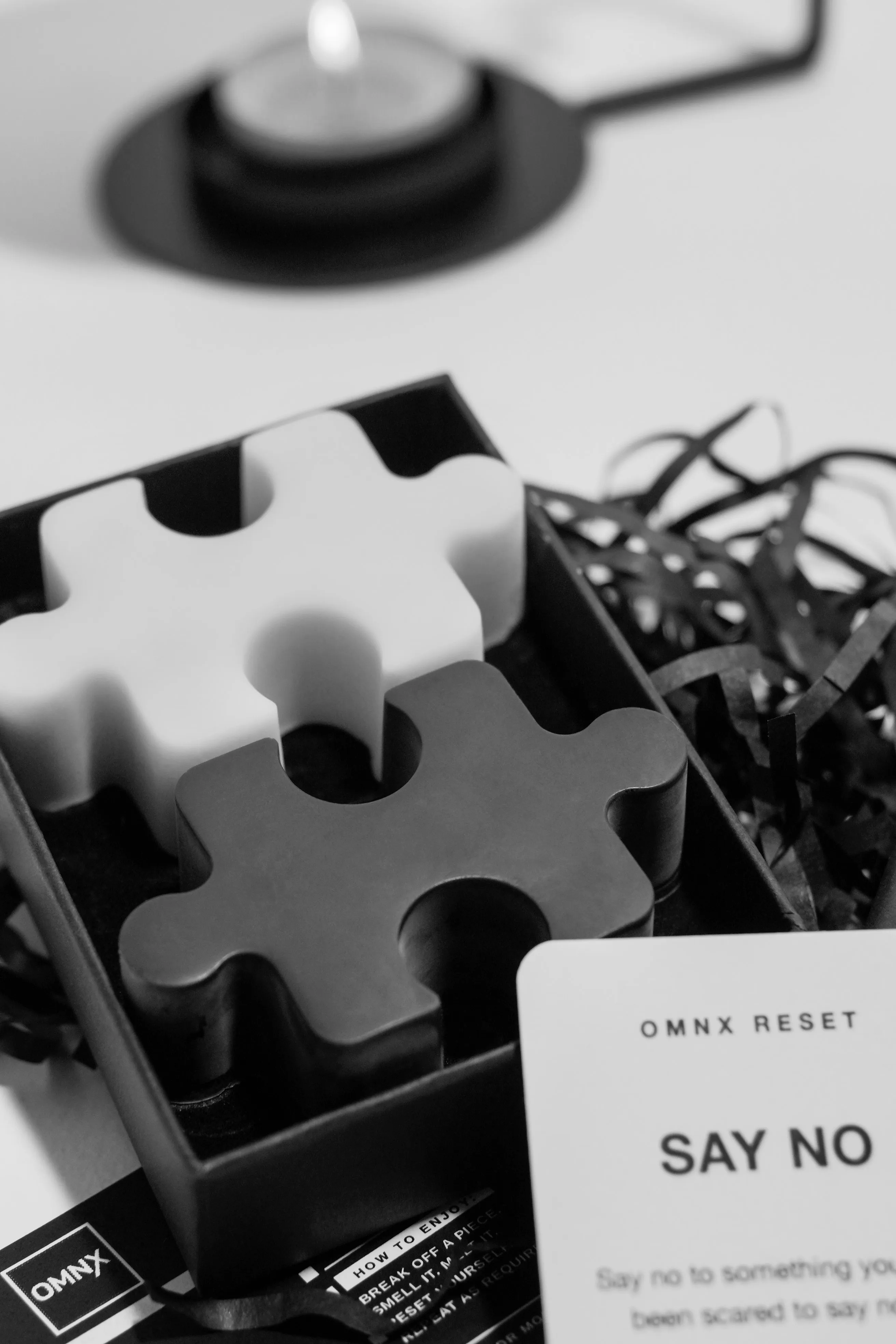 The two Noir pieces in their box, the black one nearer the camera and overlapping the white one, a lit tea light on its burner behind and an OMNX RESET card in front