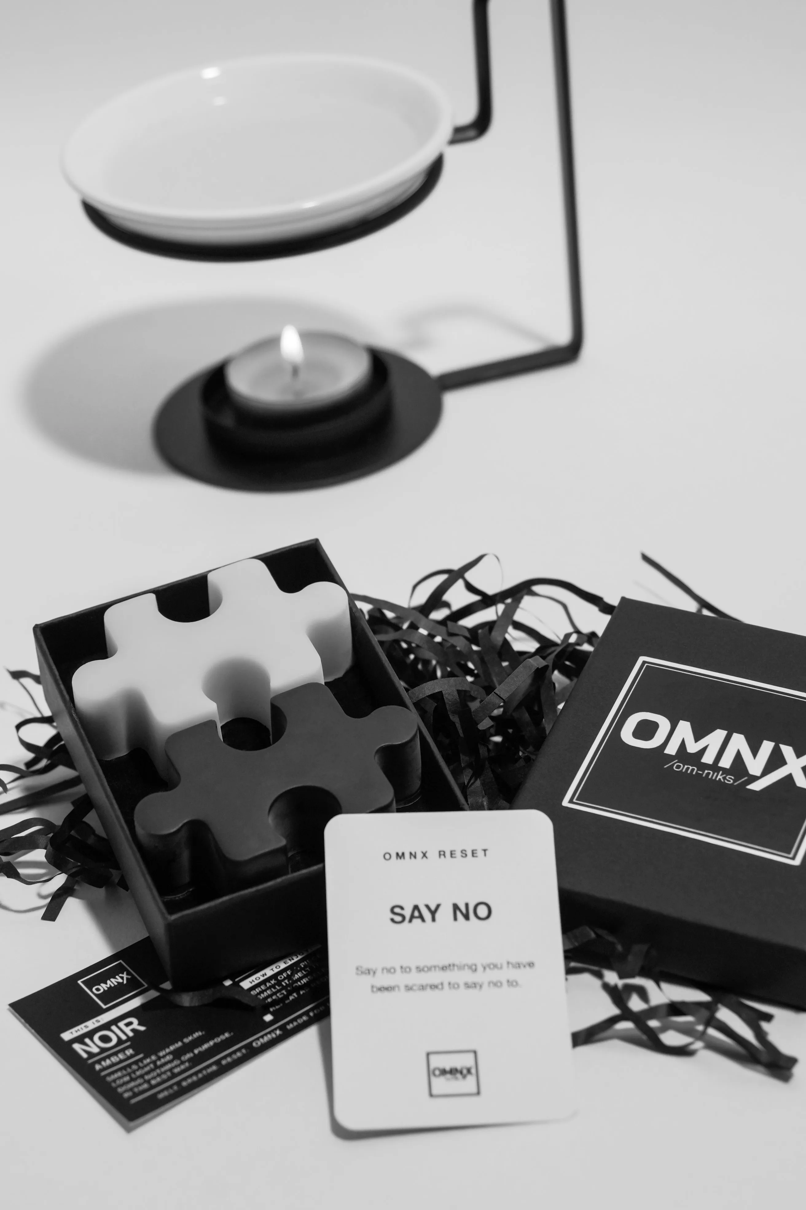 A black and white photograph of a ceramic burner, its dish empty and a tea light burning beneath it, set behind an open Noir box holding one white and one black interlocking piece, with the OMNX lid, the Noir scent card and an OMNX RESET card in front