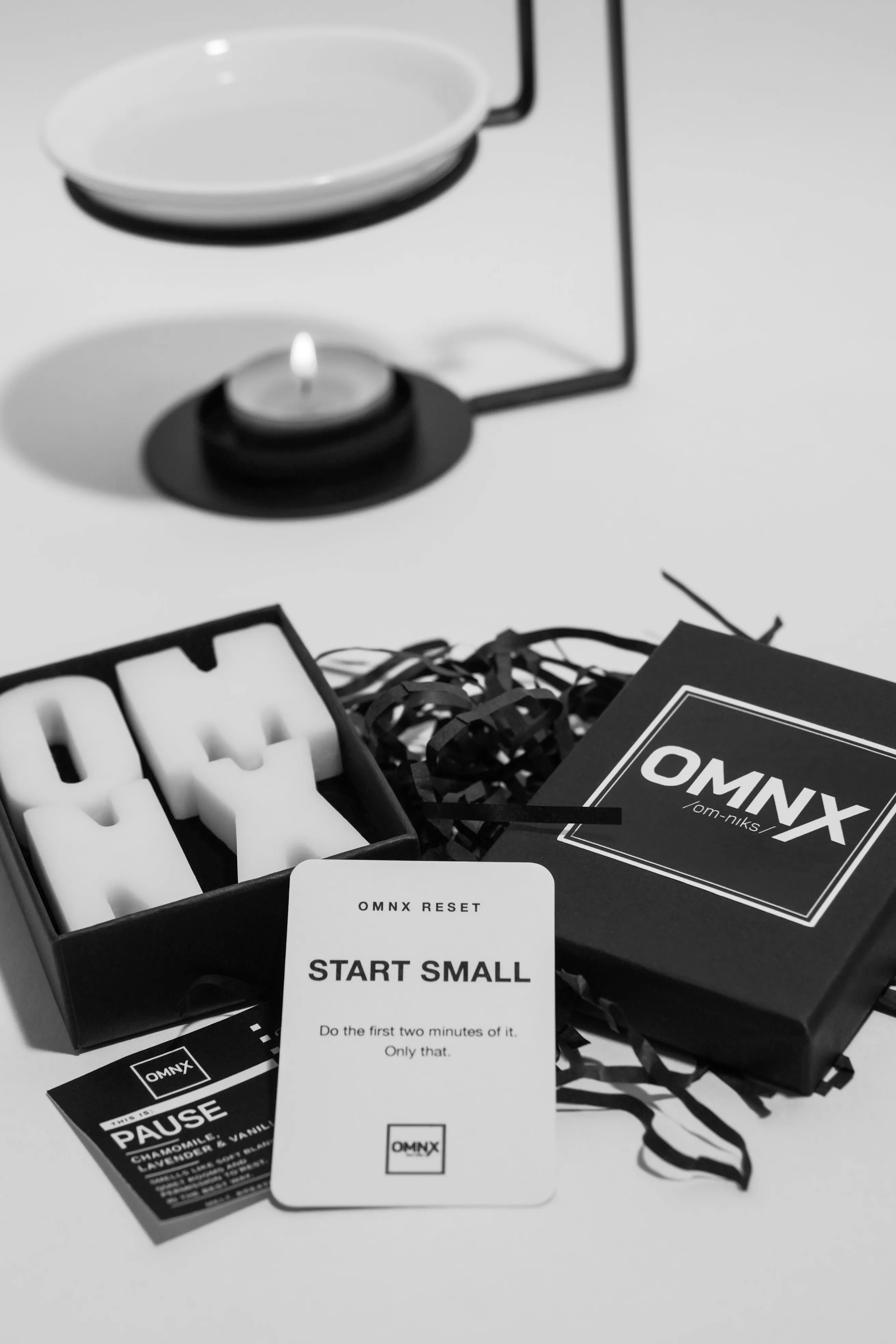 A black and white photograph of a ceramic burner, its dish empty and a tea light burning beneath it, set behind an open Pause box of pale wax letters spelling OMNX, with the lid, the Pause scent card and an OMNX RESET card in front
